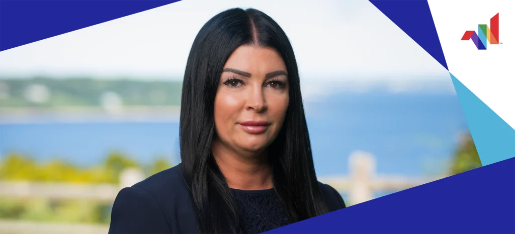 Woman with long dark hair in a navy blazer stands outdoors with a blurred background of water and greenery; geometric blue and white shapes frame the image, with a multicolor logo in the corner.