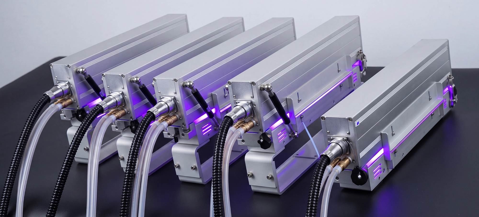 Five industrial metallic devices with attached tubes and cables are arranged in a row on a black surface, illuminated by purple LED lights.