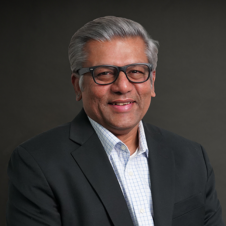 Smiling older man with gray hair and glasses wearing a dark suit jacket over a light checkered shirt against a plain dark background.