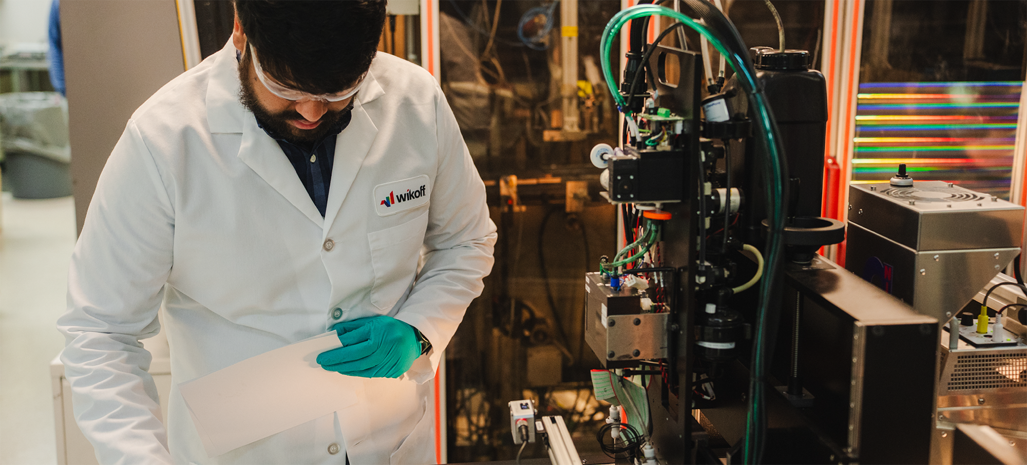 A person in a lab coat and gloves examines a sheet of material next to scientific equipment in a laboratory setting.