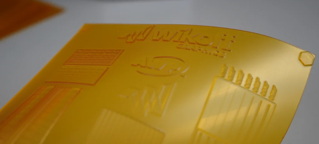 A close-up of a yellow printing plate with embossed logos, text, and patterns, placed on a white surface.