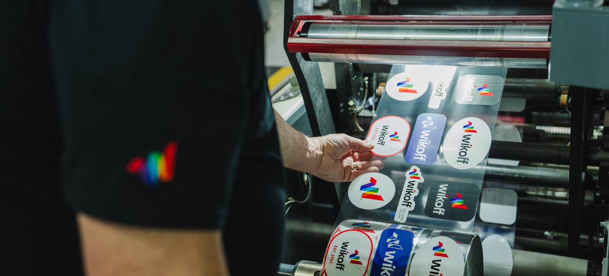 A person inspects a roll of printed labels with the Wikoff logo on a machine in a printing facility.